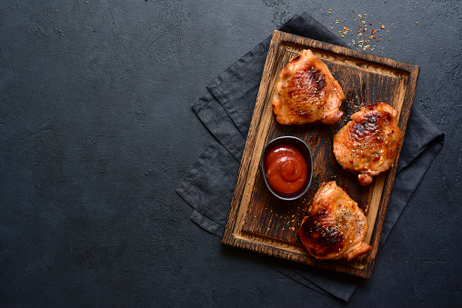 Roasted chicken thighs with ketchup on a rustic wooden board over black slate, stone or concrete background. Top view with copy space.