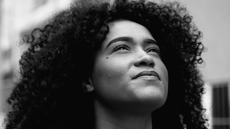 One contemplative young black woman close-up face looks up at sky feeling inspired and uplifted by TRANSCENDENCE and GODLY empowerment. African American feeling GRACE in black and white