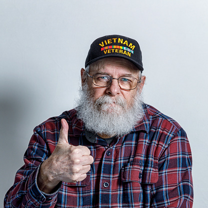 Portrait of a real life, real person senior adult man United States Navy USA Vietnam War military veteran with a full, bushy, gray beard. He is wearing eyeglasses and looking at the camera with a friendly facial expression while giving a confident, supportive thumbs up hand gesture. He's wearing a commemorative Vietnam War Veteran's baseball cap style hat.