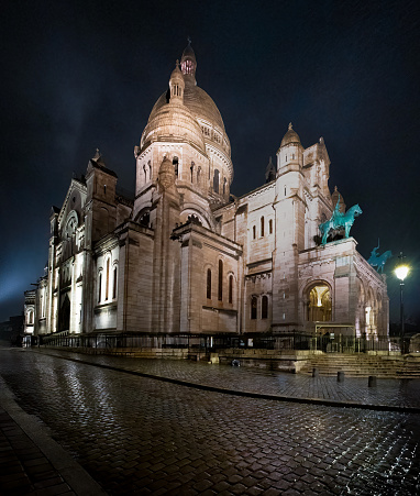 The famous Parisian landmark, the Basilica of Sacre Coeur shot on a rainy night with reflections in the wet cobblestone streets. There is some moody mist in the air