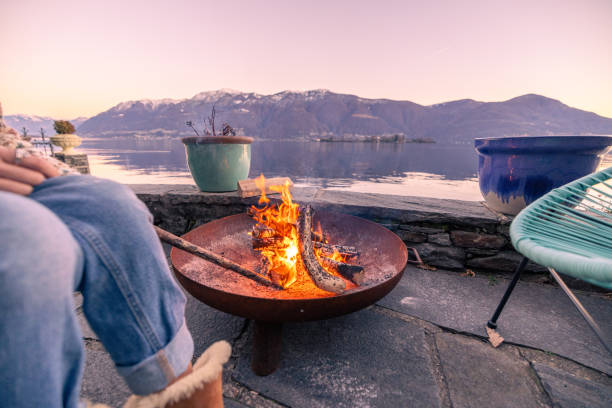 Woman enjoys outdoor fire at her home rental, stunning lake and mountain view Embracing staycations in Europe, Enjoying an outdoor moment by the fire, vacation rental, staycation concept Ultimate Cozy Atmosphere with Your Fire Pit stock pictures, royalty-free photos & images