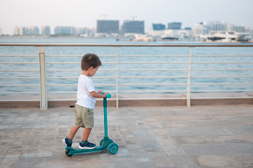 Experience pure delight as a charming two and a half year old multiracial boy cruises along the promenade by the sea on his vibrant blue scooter. Heartwarming scene of innocence and joy against the scenic backdrop of the seaside