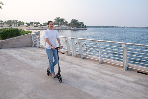 Witness the confidence and style of a young Chinese woman as she cruises along the seaside promenade in jeans and a t-shirt, embracing the coastal breeze on her scooter. A blend of fashion and freedom against the picturesque backdrop of the sea