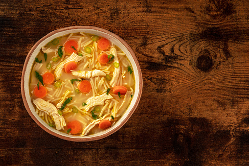 Chicken noodle soup with vegetables, a bowl of healthy broth, shot from the top on a rustic wooden table, winter comfort food with a place for text