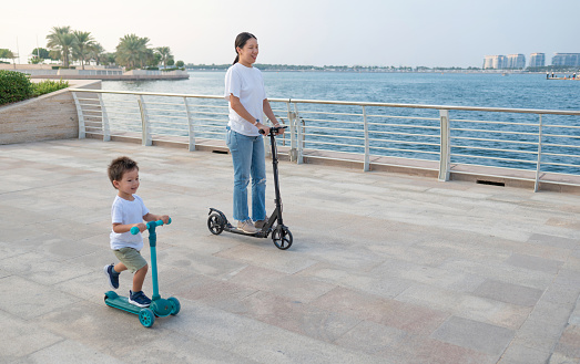 Delight in the seaside breeze as a charming two and a half year old multiracial boy and his Chinese mother enjoy a carefree scooter ride along the scenic promenade. A heartwarming blend of cultural diversity and mother-son joy against the backdrop of the serene sea