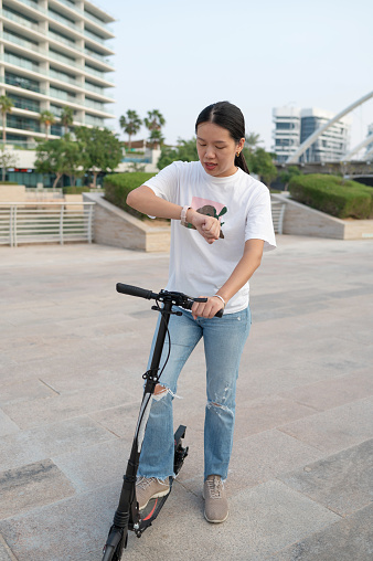 Embrace the fusion of style and punctuality as a young Chinese woman in jeans and a T-shirt, riding a scooter, gracefully checks the time on her wristwatch. A moment of urban chic captured in the midst of a fashionable and timely scooter adventure