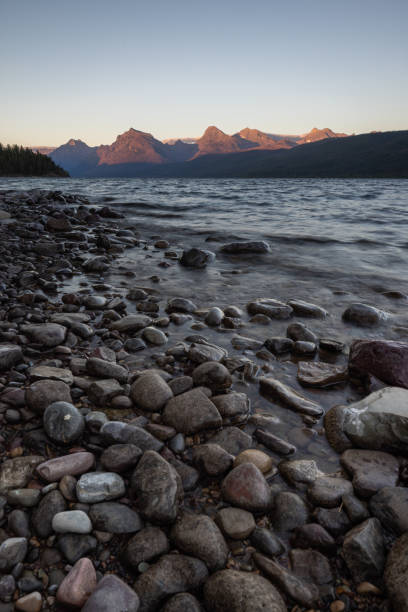 A calm autumn morning at Lake McDonald in Glacier National Park, Montana. The clear water reveals colorful stones beneath the surface while mountains and trees reflect in perfect symmetry across the still lake.