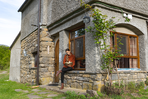 Woman with crossed legs reads at the entrance of a country house, concentrated, turns the page