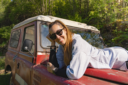 Woman smiling, lying on the hood of an old red off-road vehicle parked in the middle of the Pyrenees forest