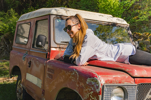 Woman lying on the hood of an old red off-road vehicle parked in the middle of the Pyrenees forest very happy and smiling