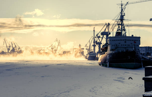 Big ship on snowy frozen river surrounded by construction cranes in background, beautiful foggy sunset