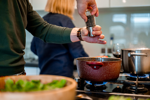 Hands of adult man adding some pepper or other spice into a red cooking pan in a domestic kitchen. Unrecognizable woman in the background.