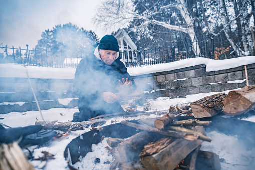 Young man starting outdoor fire at the fire pit in winter. He is  dressed in warm winter outdoor clothing. Exterior of Lake House in Georgian Bay area of Ontario, Canada.