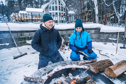 Young Couple starting outdoor fire at the fire pit in winter. Both dressed in warm winter outdoor clothing. Exterior of Lake House in Georgian Bay area of Ontario, Canada.