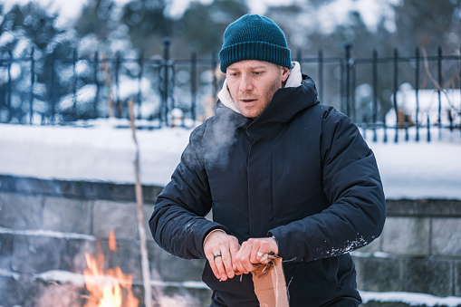 Young man starting outdoor fire at the fire pit in winter. He is  dressed in warm winter outdoor clothing. Exterior of Lake House in Georgian Bay area of Ontario, Canada.