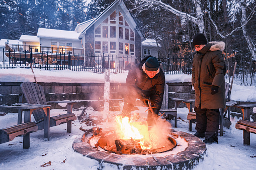 Family members starting outdoor fire at the fire pit in winter. Both dressed in warm winter outdoor clothing. Exterior of Lake House in Georgian Bay area of Ontario, Canada.