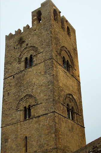 Close-up view upper part of stone belltower of ancient Erice Cathedral in small mountain village Erice. Notable landmark of Sicily. Travel and tourism concept. Cloudy sky background.