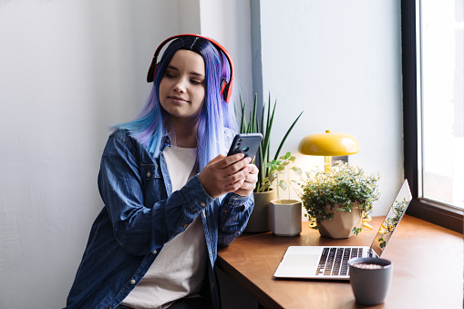Portrait of attractive young teenager latin hispanic girl with blue hair and blue denim shirt sitting in cafe and using smartphone, with copy space.