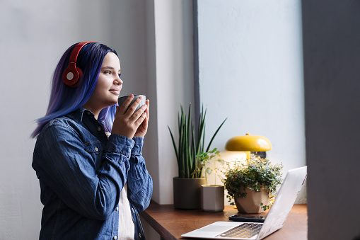 Portrait of attractive young teenager latin hispanic girl with blue hair and blue denim shirt sitting in cafe and drinking hot tea or coffee, with copy space.
