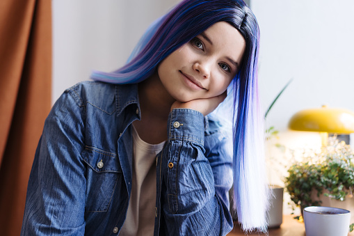 Portrait of attractive young teenager latin hispanic girl with blue hair and blue denim shirt sitting in cafe and looking at camera, with copy space.