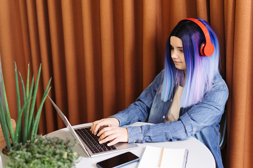 Portrait of attractive young teenager latin hispanic girl with blue hair and blue denim shirt sitting in cafe and typing on a laptop, with copy space.