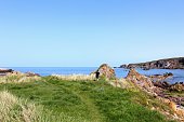 A man walks along the scenic rocky coast of the highlands in Scotland, near Portknockie.  The beautiful ocean and rocky shoreline shimmers on a sunny summer day with blue sky.
