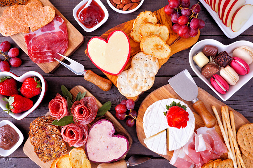 Valentine's Day theme charcuterie table scene against a dark wood background. Variety of cheese, meat, fruit and sweet appetizers. Overhead view.