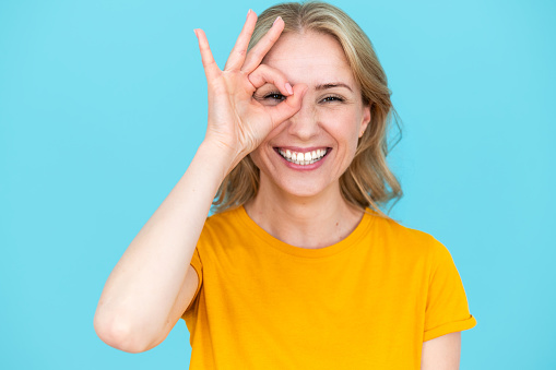 Portrait of smiling young woman peeping through ok gesture over blue background. Happy female winking at camera and smiling. Overjoyed, positive emotions concept