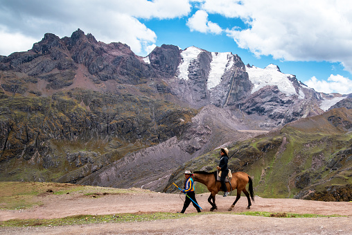 A Quechuan man guiding a tourist on a horse on Rainbow Mountain trail. \n The trail has a peak elevation of over 17,000 feet.