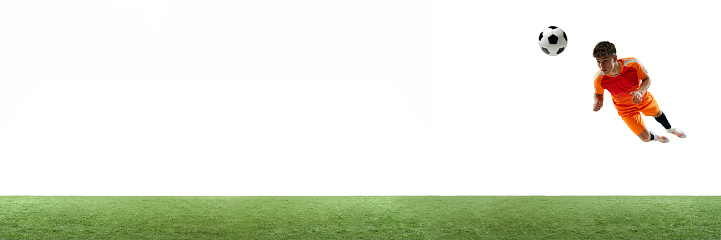 Young man, football player in orange hitting ball in a jump with head isolated over white background with grass flooring. Concept of sport, game, competition, championship, active lifestyle. Banner