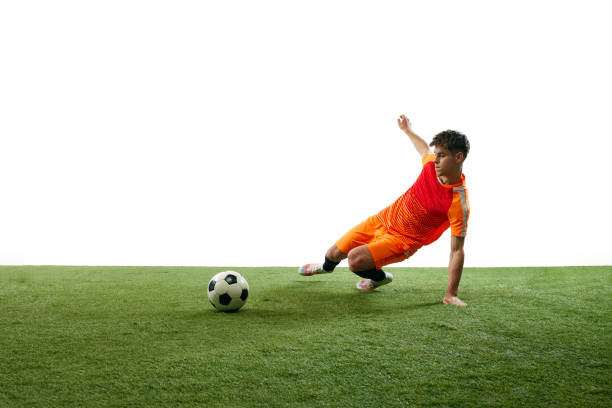 Competitive young man, soccer playing in motion during game, falling down, hitting ball isolated over white background with grass flooring. Concept of sport, game, competition, active lifestyle