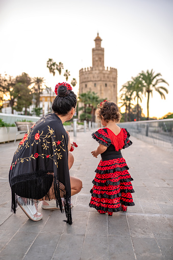 Rear view of mother and young daughter dressed in red flamenco dress in front of the Torre del Oro in the morning, Seville, Spain