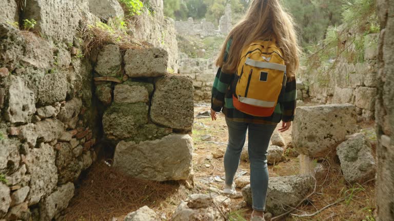 The camera follows a woman with a backpack as she navigates through a narrow and dark tunnel among the stones of an ancient city, emerging into the sunlight in the forest. Archaeological journey.