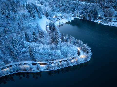 Aerial view of road, snowy forest, illumination, lake, street lights at winter night. Top drone view of alpine countryside, road through the snowy pine trees at dusk. Bled lake, Slovenia. Travel
