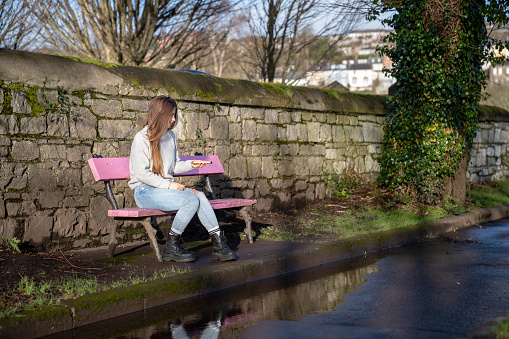 White, brown-haired woman wearing a gray sweater, jeans and black boots, sitting on a pink bench on a sunny morning in Ireland