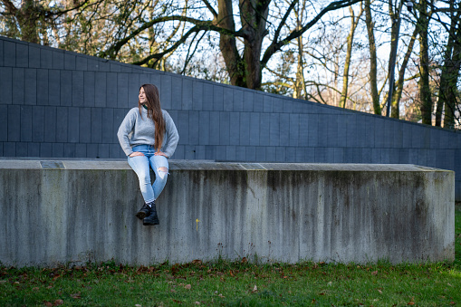 White, brown-haired woman wearing a gray sweater, jeans and black boots, sitting on a pink bench on a sunny morning in Ireland