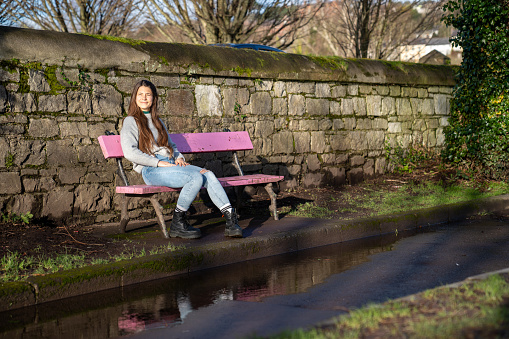 White, brown-haired woman wearing a gray sweater, jeans and black boots, sitting on a pink bench on a sunny morning in Ireland