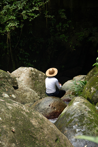 young person with white shirt and hat sitting on a rock in the outdoors by the river