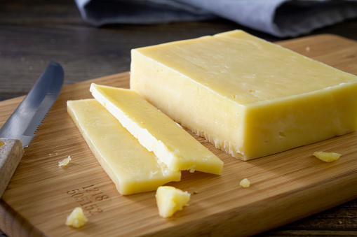 A block of cheddar cheese cut into slices on a chopping board. On a rustic wooden kitchen table kitchen utensils and herbs in the background.
