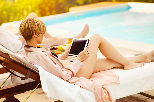 Beautiful happy couple lying on loungers by the pool and using tablet and smart phone devices
