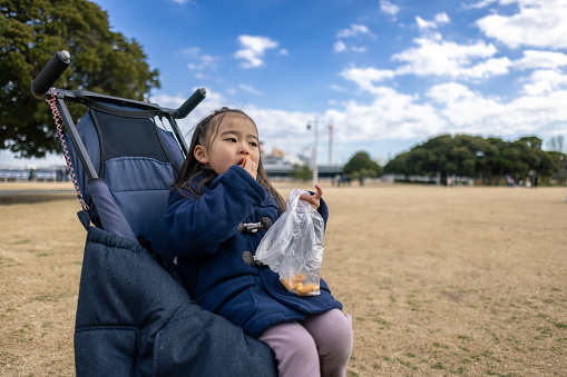 Little girl sitting on baby carriage and eating snacks in public park