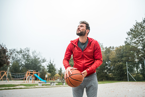 A Hispanic male player in red hoodie, shooting a basketball outside.