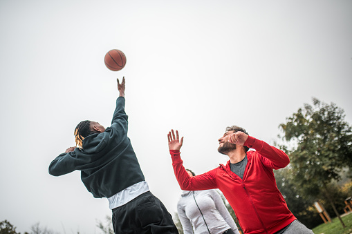 A competitive jump ball situation featuring a Black male and a Hispanic male in an outdoor basketball game.