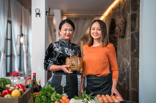 Portraits of beautiful Chinese mature female chefs dressed in traditional black Chinese Cheongsam and modern orange top posing in front of the camera in the restaurant with fresh ingredients on the checked patterned table.