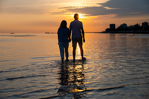 Senior adult couple silhouettes holding hands and walking to sea barefoot at sunset, wide shot back view