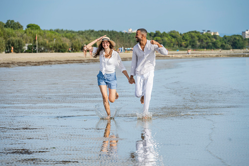 Happy young multiracial couple holding hands and running along coastline, wide shot