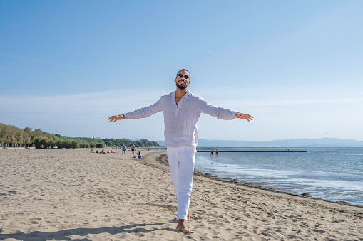 Smiling handsome multiracial man spreading hands while walking barefoot on beach and looking at camera, full length portrait