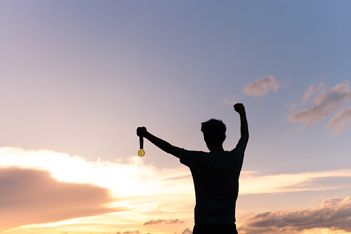 Silhouette of an athlete celebrating the gold medal against a sunset sky adorned with golden clouds in the background. Ideal for conveying the spirit of success in sporting events.