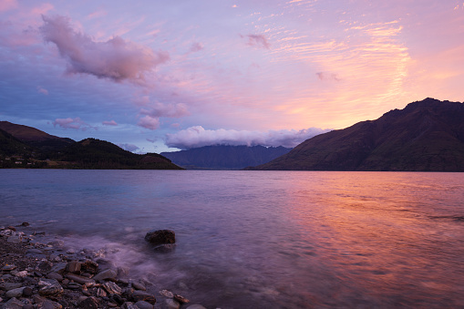 Sunrise at Lake Wakatipu, taken from 12 Mile Delta, just a short distance away from Queenstown, New Zealand. The colorful sunrise reflects in the water, while a gentle wave breaks on the shore. In the distance, with a cap of while cloud is the well-known Remarkables mountain range.