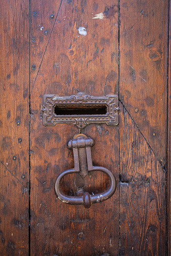 A tarnished antique metal door knocker and mail slot on a dark brown, weathered wood panel door.